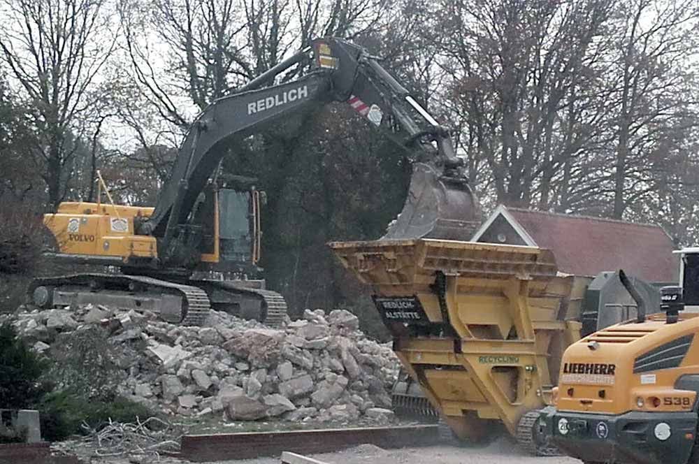 Ein Bagger verlädt Schutt auf einer Abrissbaustelle in einen gelben Brecher.