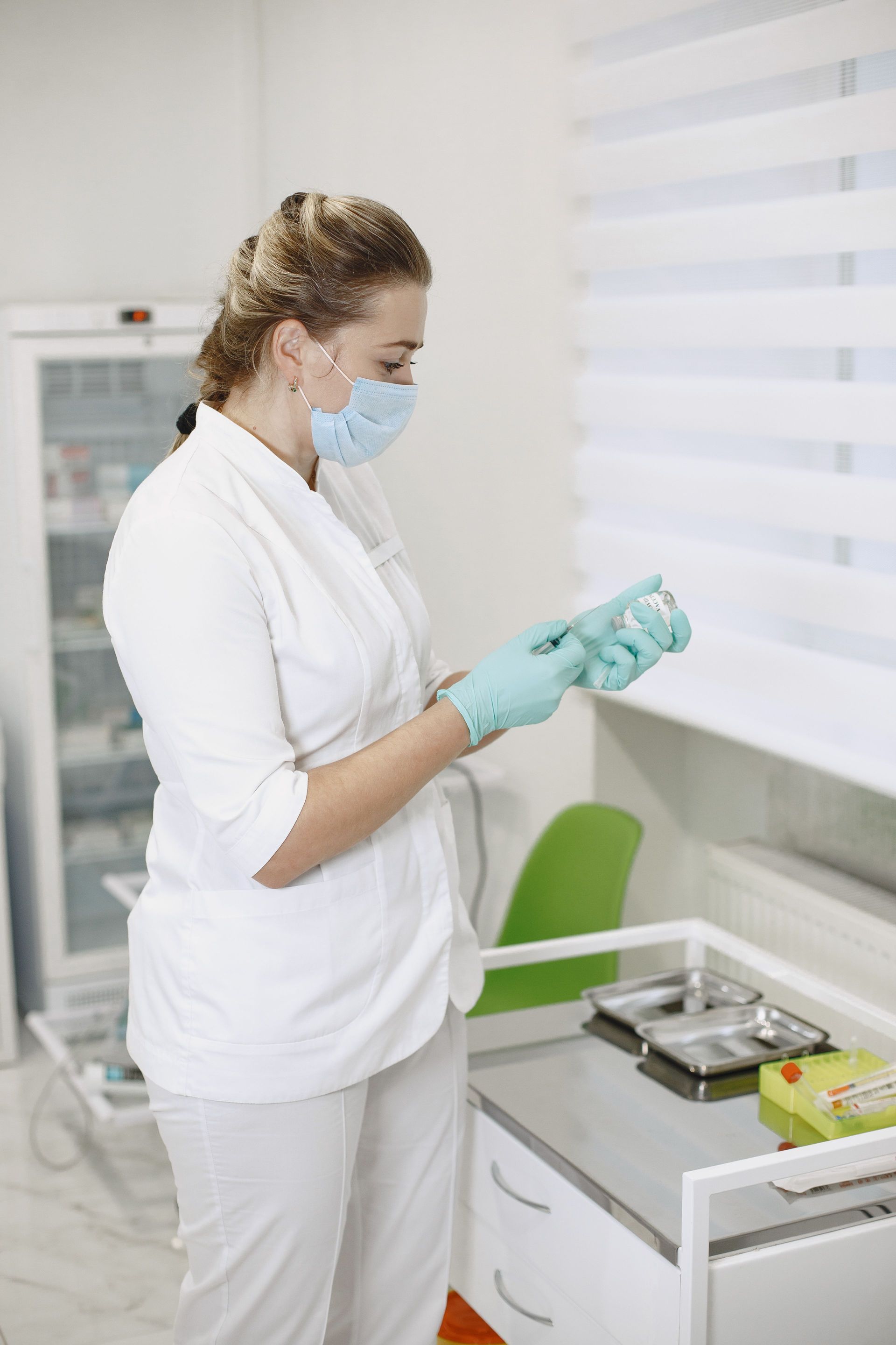 Enfermera con uniforme blanco y guantes preparando medicamentos junto a una ventana y un carrito del hospital