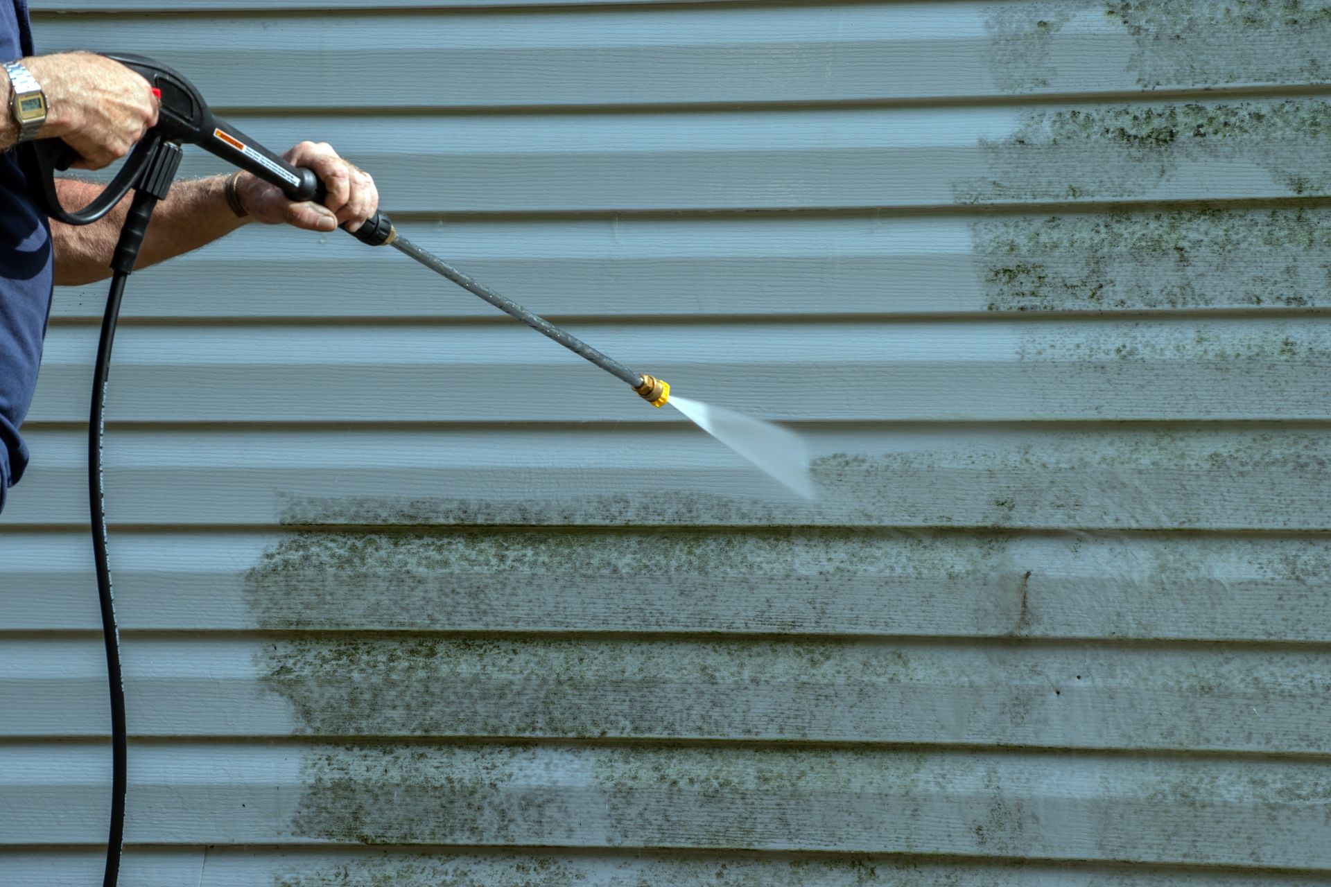 Une personne nettoie à haute pression un mur de bardage gris avec un jet d'eau.