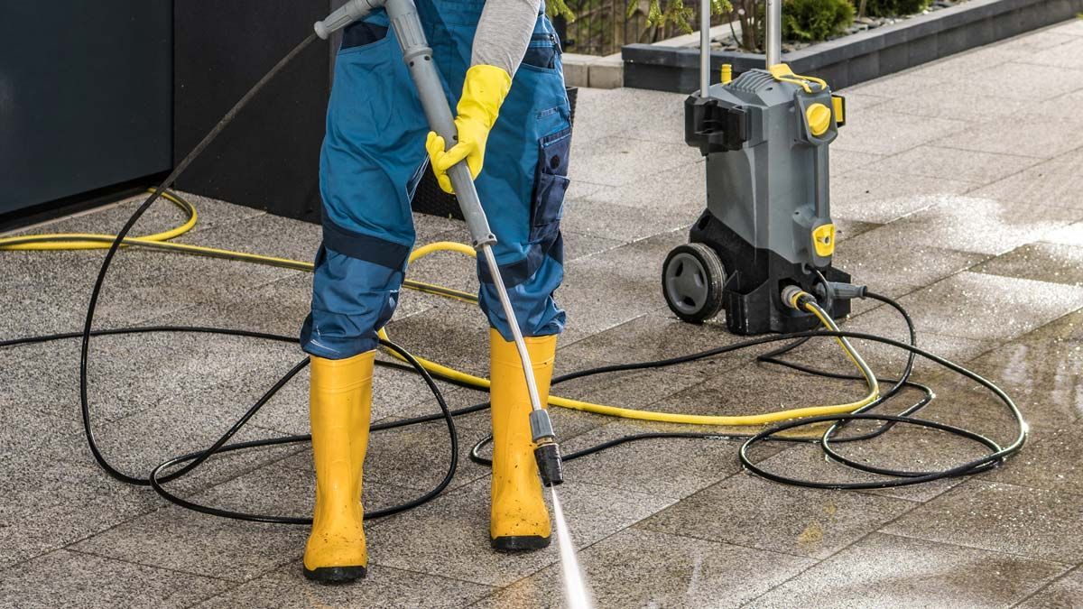 Un ouvrier en bottes jaunes nettoie à haute pression une surface en béton humide à côté d'une machine de nettoyage sur roues.