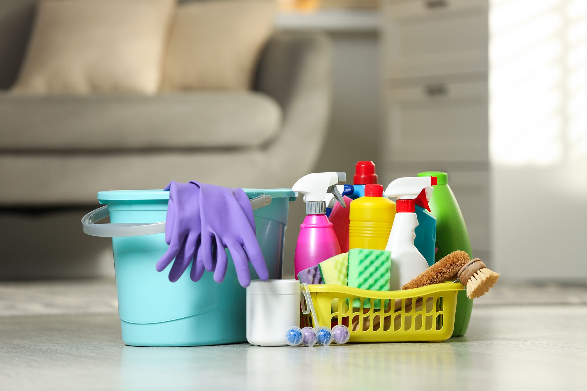 Des produits de nettoyage dans des contenants colorés, disposés sur le sol : gants, sprays, lingettes et brosse.