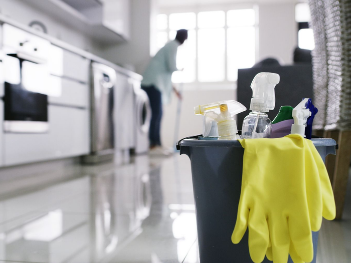 Produits de nettoyage dans une cuisine : des gants jaunes sur un seau au premier plan et une personne en train de nettoyer en arrière-plan.