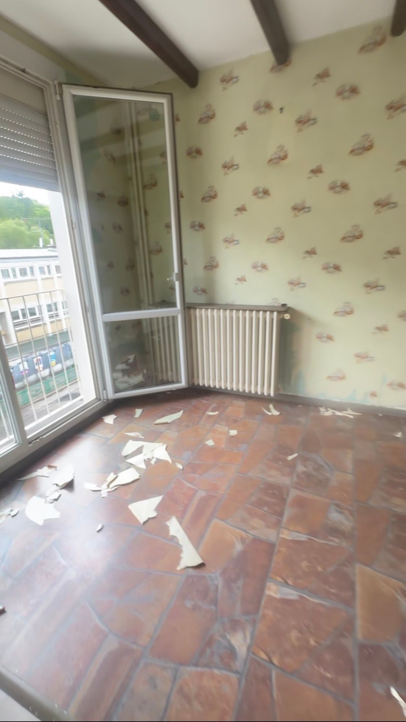 Pièce avec balcon vide, porte ouverte, sol carrelé en terre cuite, mur tapissé et débris éparpillés au sol.