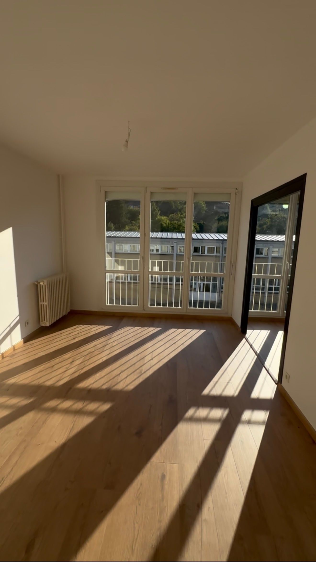 Salon lumineux et vide avec parquet, jeux d'ombres et de lumière et portes-fenêtres donnant sur un balcon.