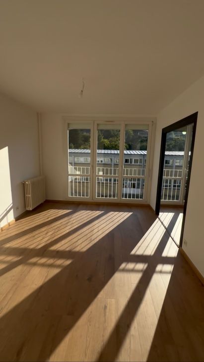 Salon vide et ensoleillé avec parquet, grandes fenêtres et longues ombres projetées par la porte-fenêtre du balcon