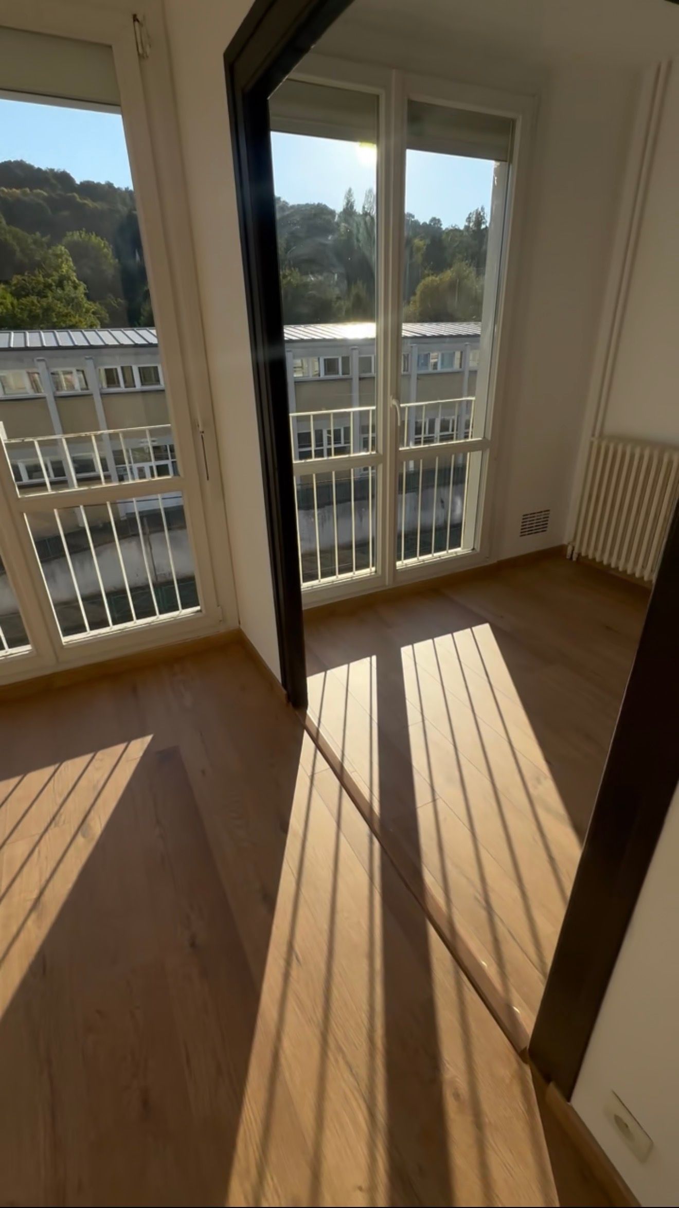 Pièce vide baignée de soleil avec portes-fenêtres donnant sur un balcon, murs blancs, parquet et longues ombres portées par les fenêtres.