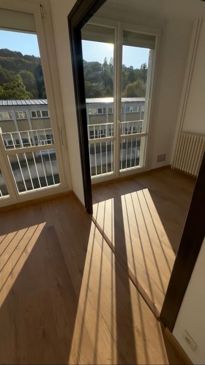 Pièce vide et ensoleillée avec portes-fenêtres donnant sur un balcon, murs blancs, parquet et longues ombres portées par les fenêtres.