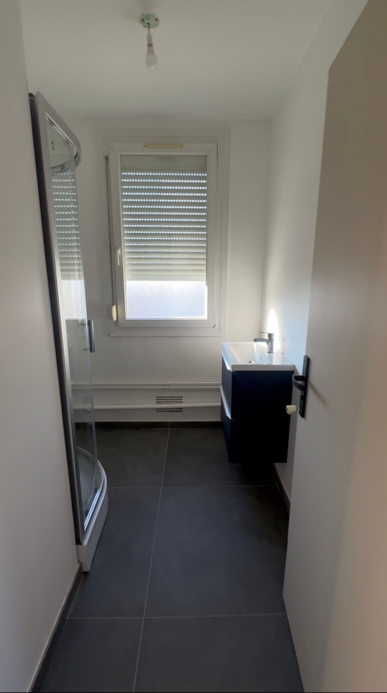 Couloir étroit et blanc avec sol carrelé, fenêtre au fond et meuble noir sur la droite.
