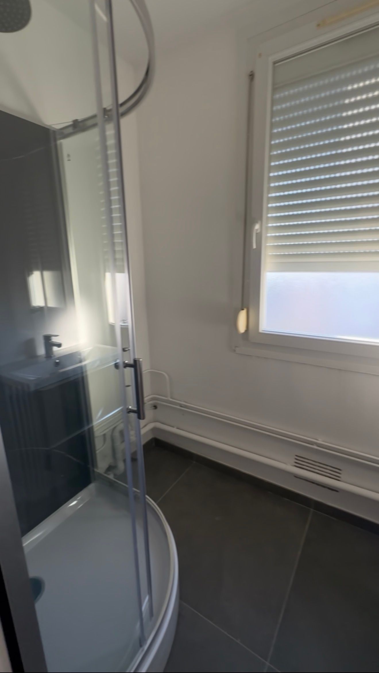 Petit coin de salle de bains avec une cabine de douche en verre, un radiateur blanc et une fenêtre avec des stores.