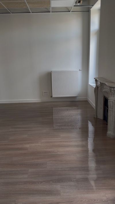 Pièce vide avec parquet ciré, murs blancs, radiateur et cheminée en briques apparentes sur la droite.
