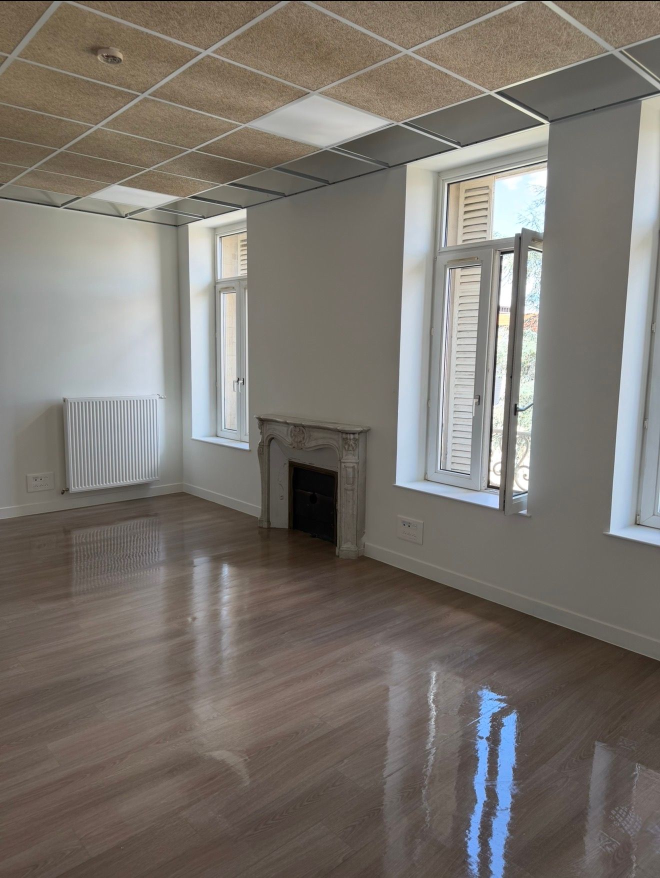 Pièce vide avec parquet brillant, murs blancs, hautes fenêtres et petite cheminée.