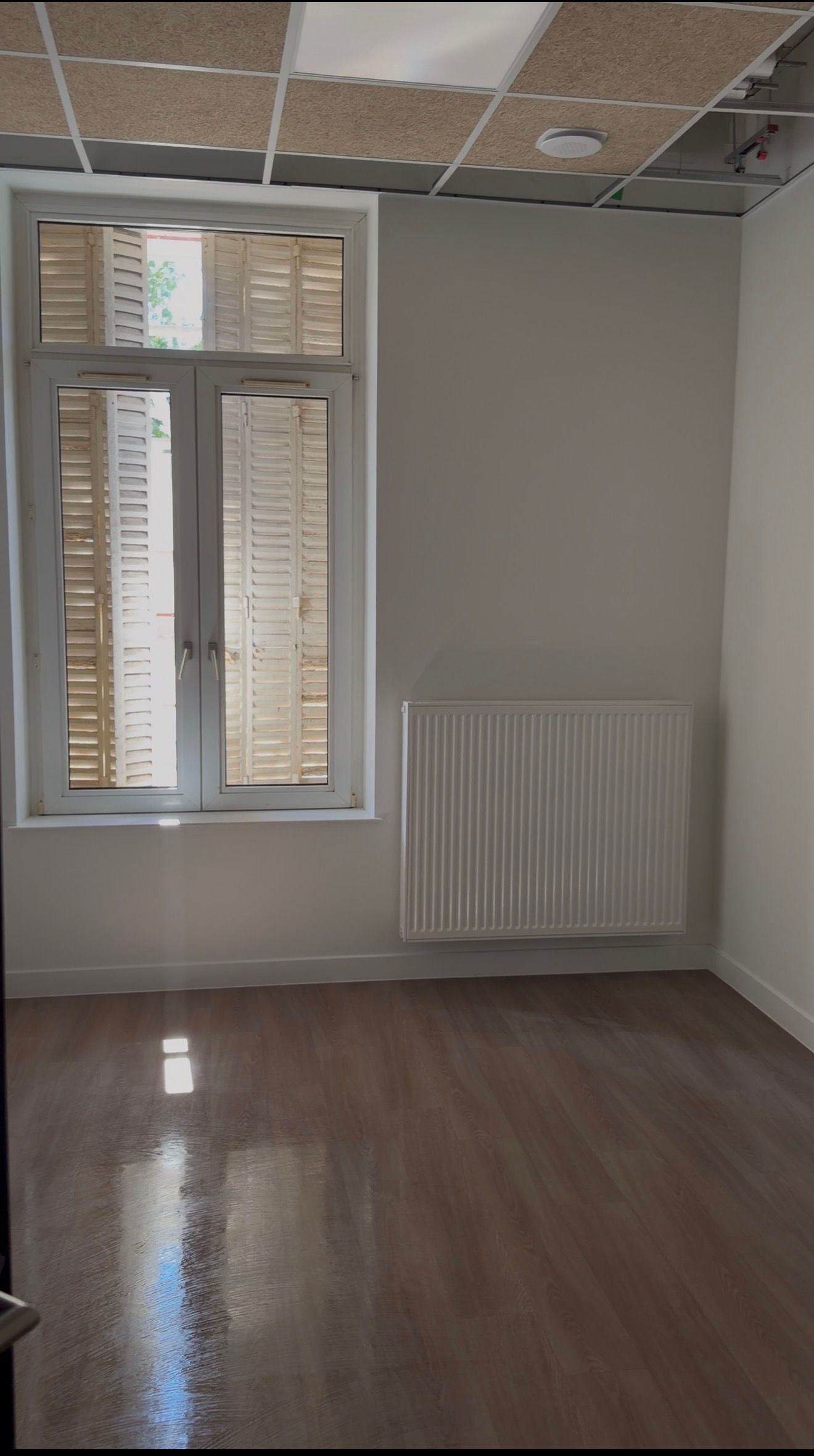 Pièce vide aux murs gris, parquet, fenêtre à volets et radiateur en dessous.