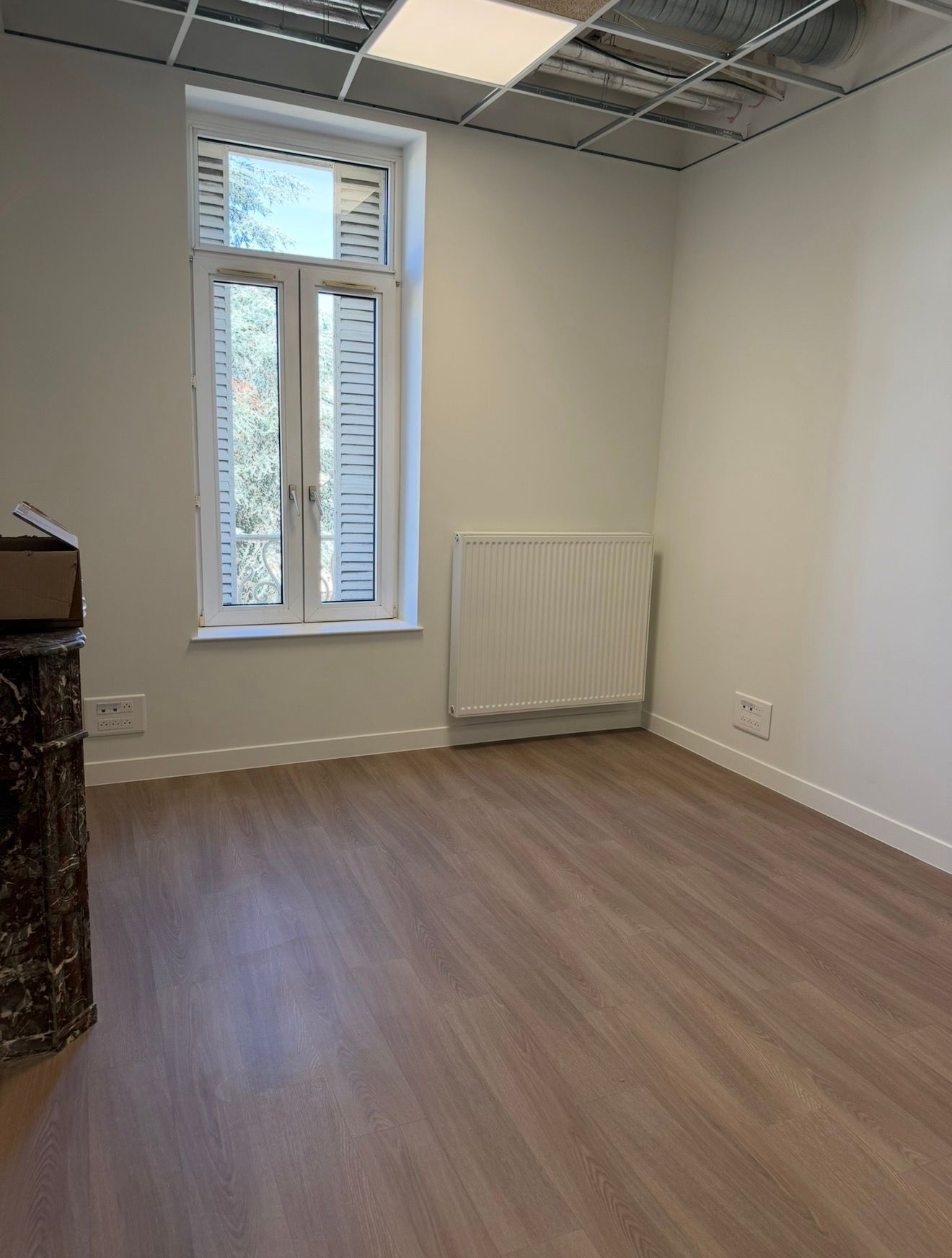 Pièce de bureau vide avec parquet, murs blancs, fenêtre, radiateur et plafonnier suspendu.