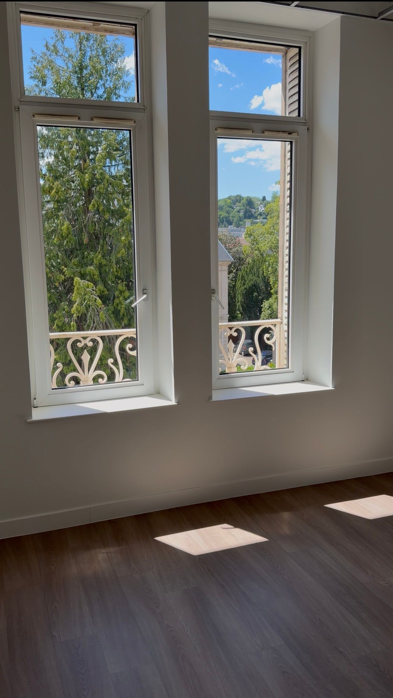 Pièce baignée de soleil avec deux hautes fenêtres, des moulures blanches et un parquet projetant un rectangle lumineux.