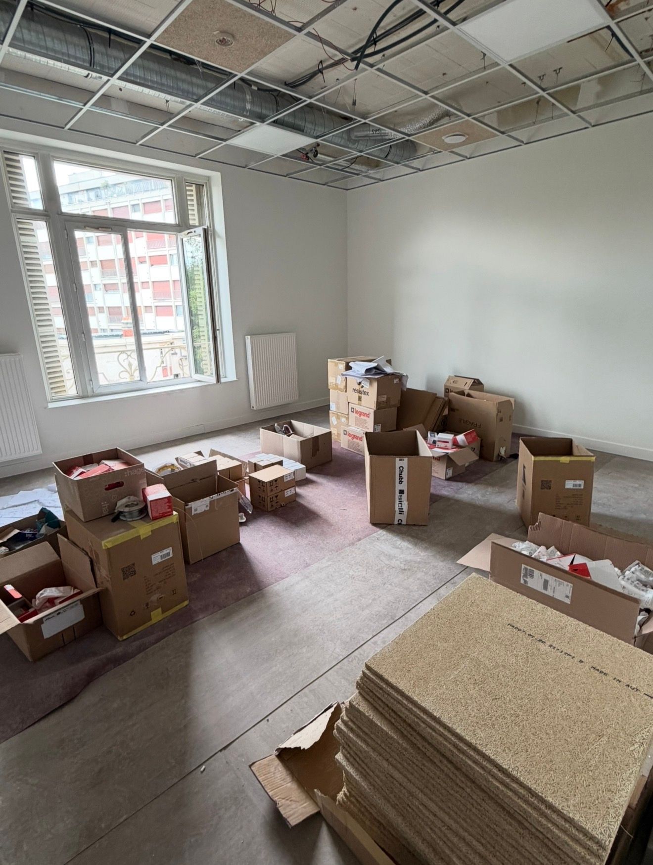 Pièce inachevée avec des cartons éparpillés, des dalles de plafond et des matériaux en kit empilés près d'une fenêtre.