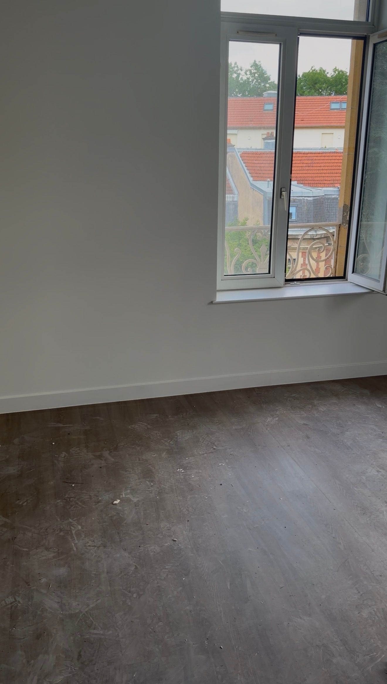 Pièce vide aux murs gris, au parquet foncé et avec une fenêtre donnant sur les bâtiments voisins.