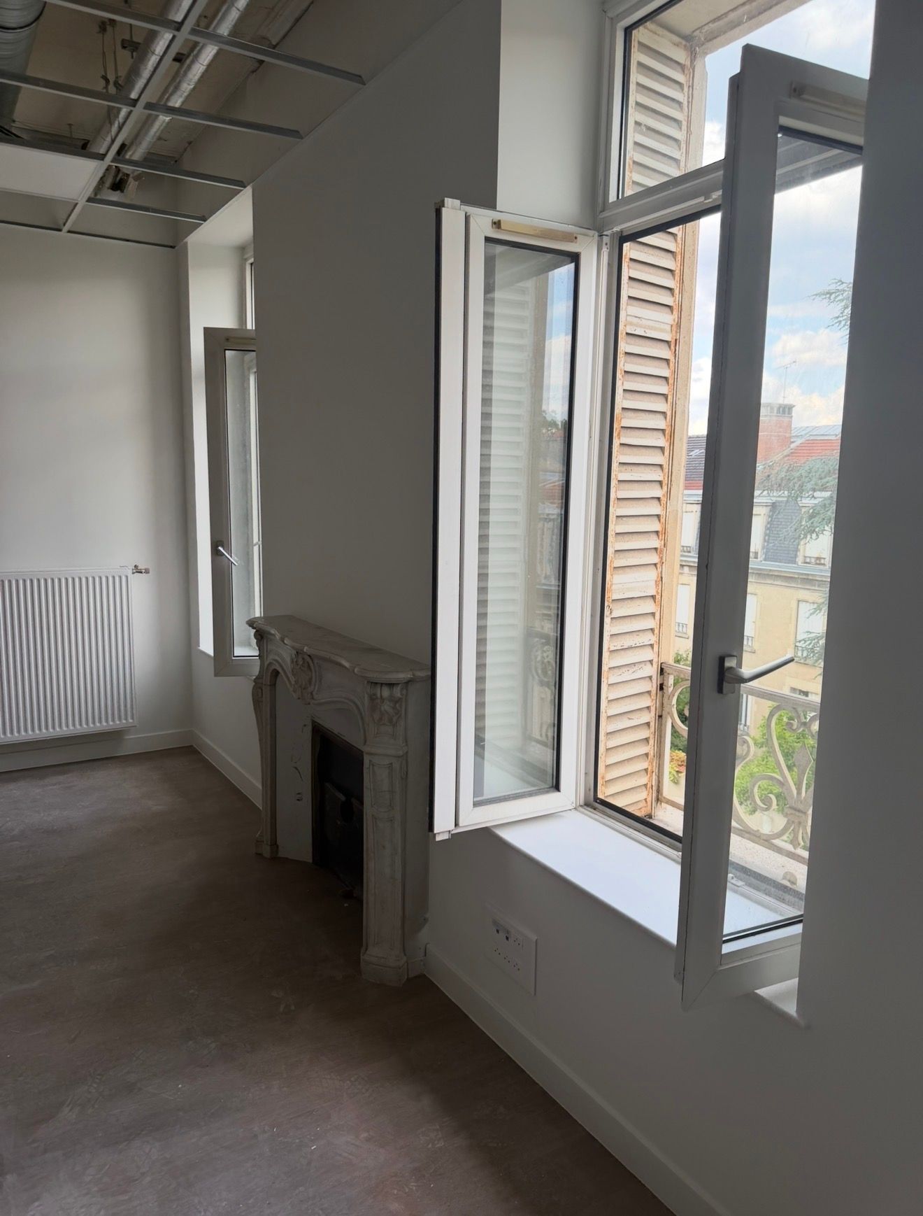 Pièce vide avec fenêtres blanches ouvertes, un radiateur et une cheminée dans un intérieur lumineux et inachevé.