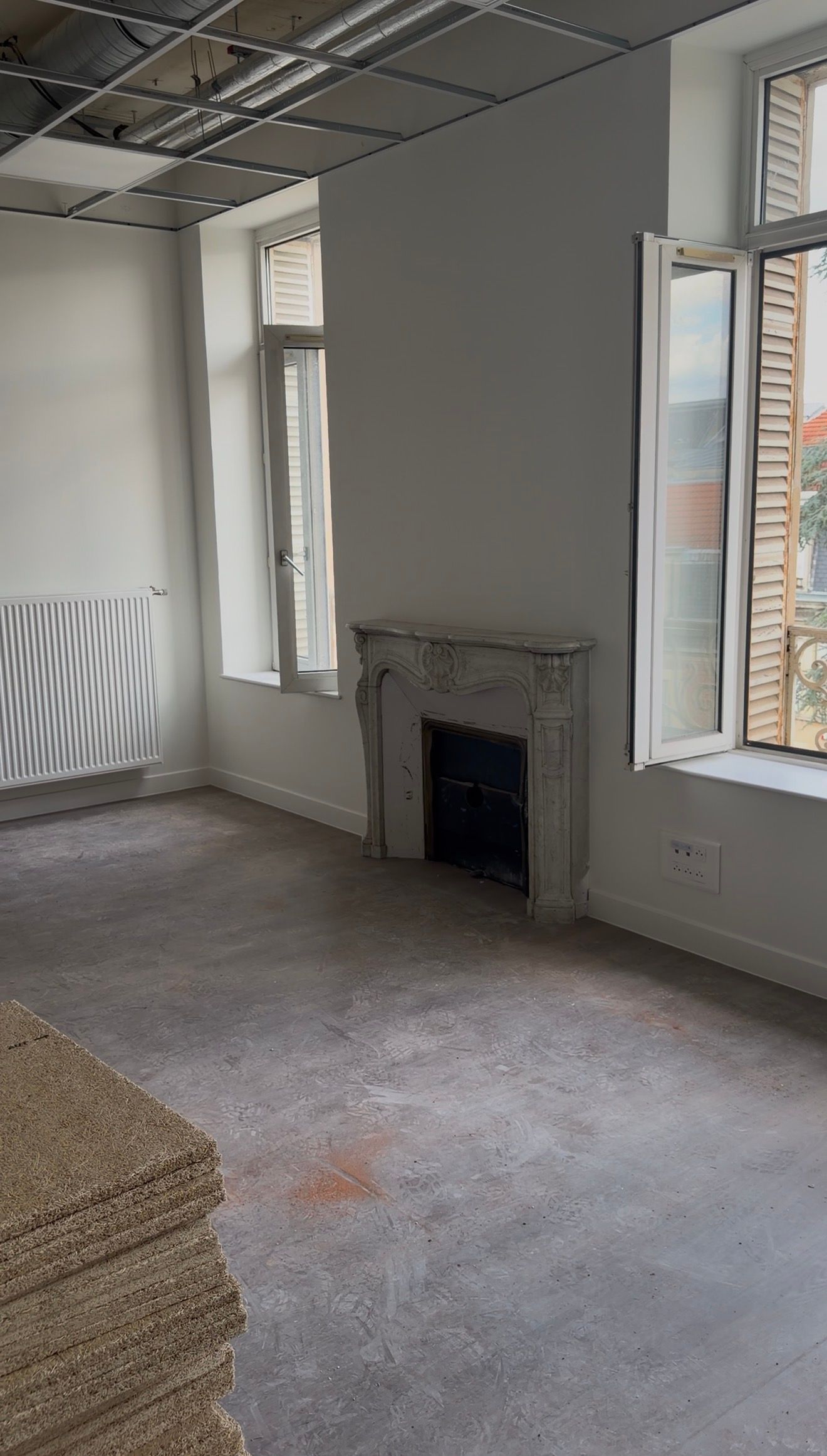 Pièce vide non meublée avec sol gris, murs blancs, hautes fenêtres et une petite alcôve avec une cheminée.