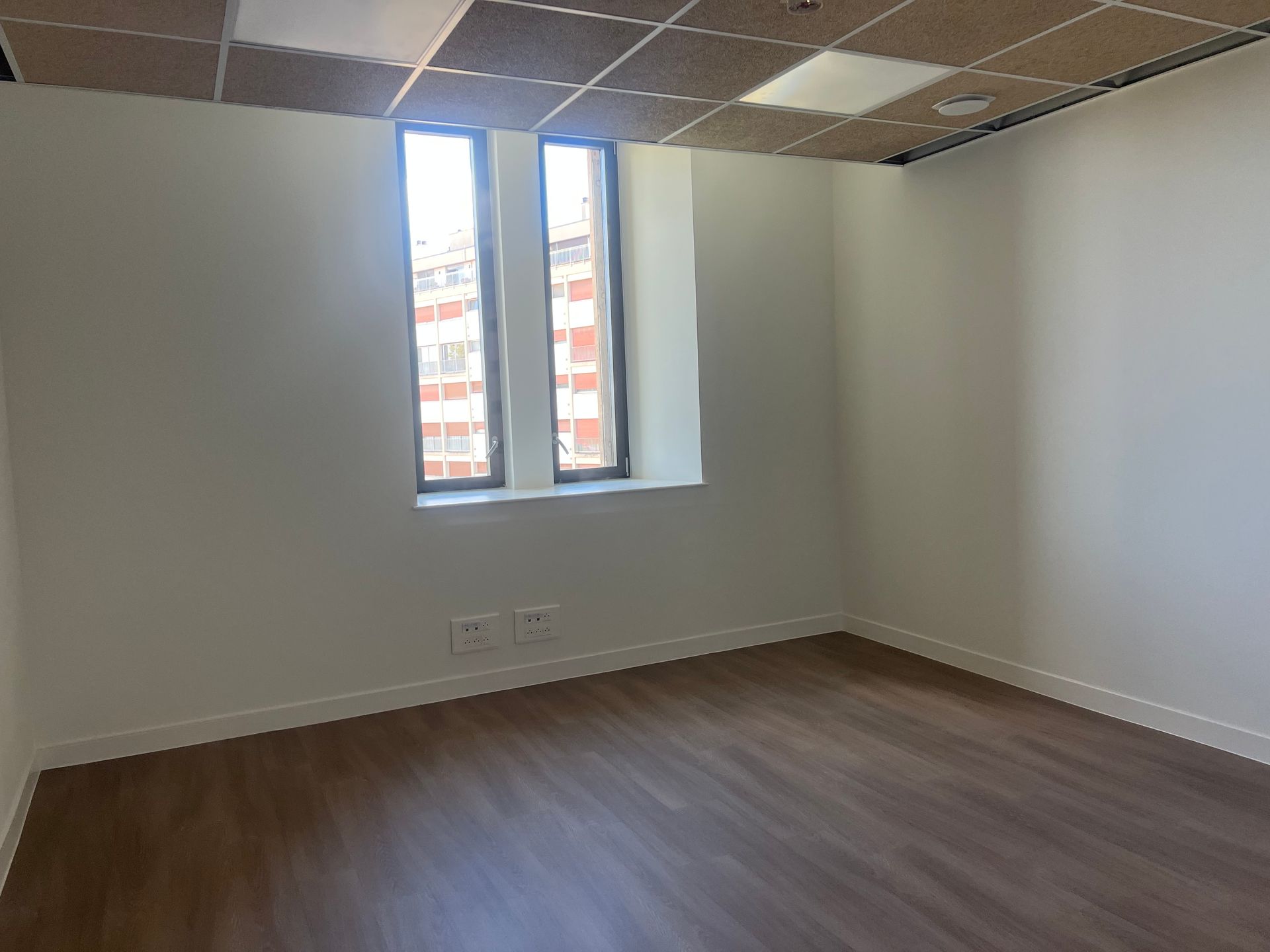 Pièce vide avec parquet marron, murs blancs, faux plafond et fenêtre donnant sur des bâtiments.