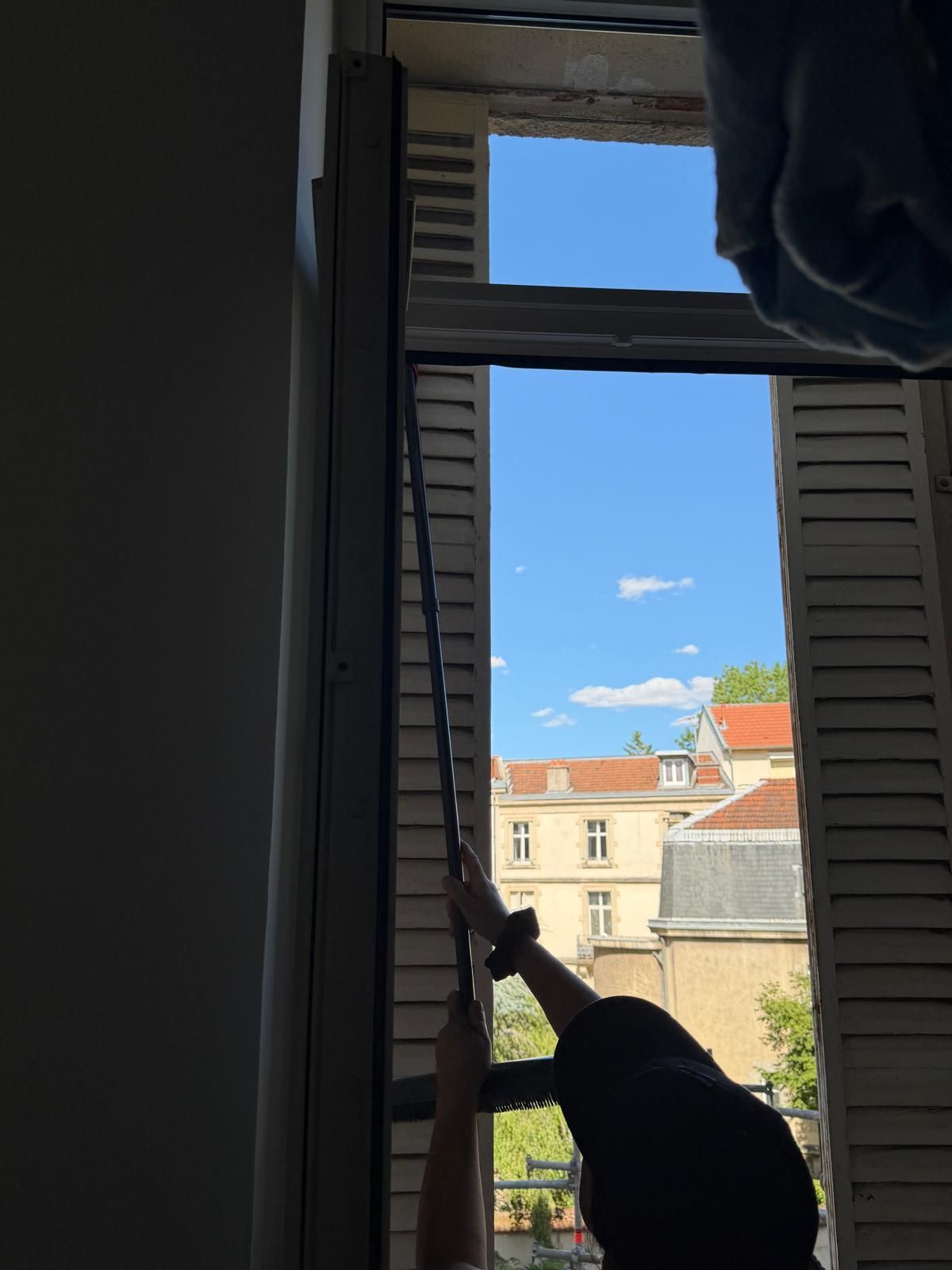 Une personne ouvre un volet sombre, regardant un ciel bleu éclatant et les bâtiments voisins.
