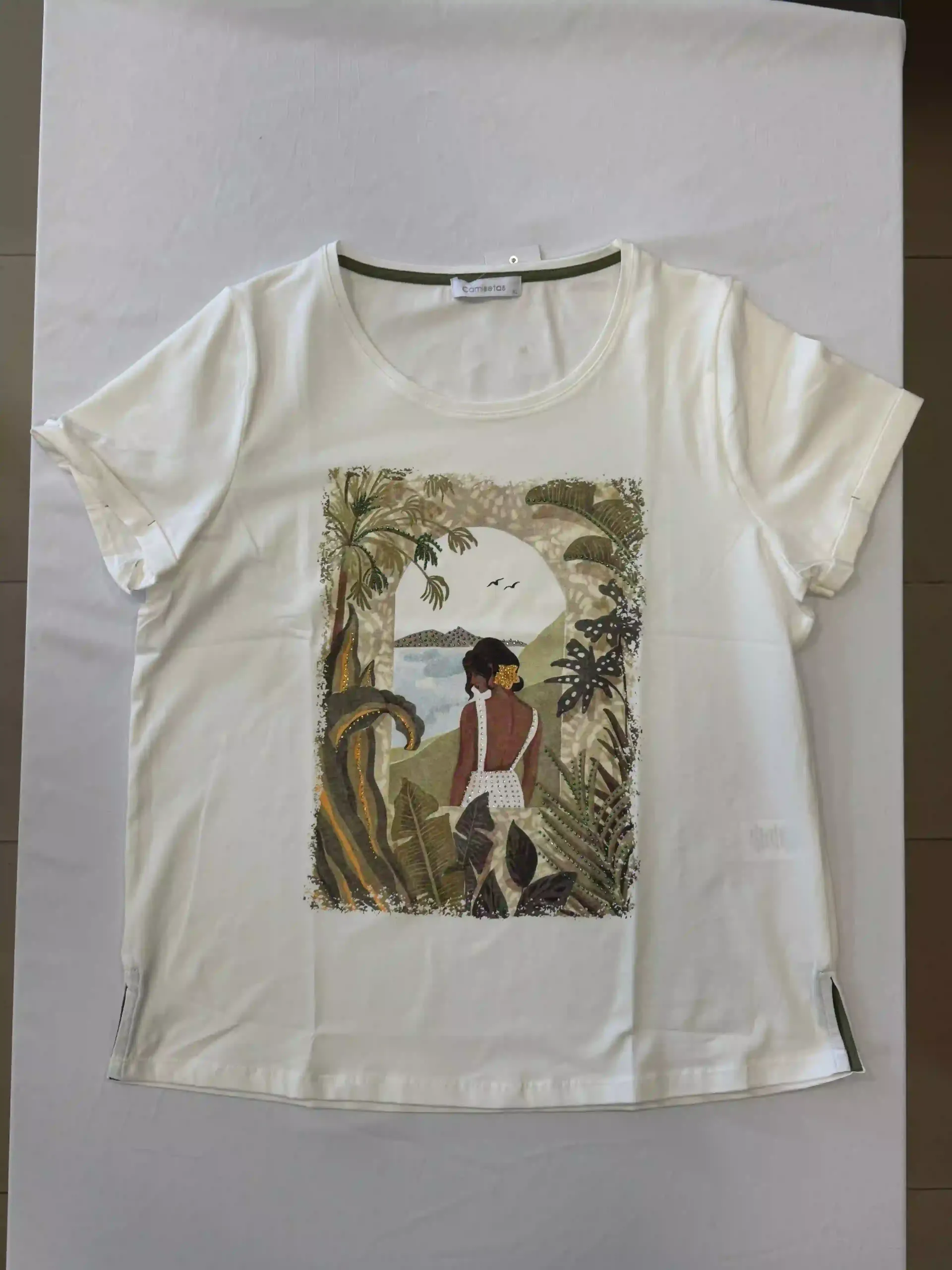 Camiseta blanca con un estampado tropical de una persona de pie entre palmeras y vegetación bajo un gran sol.