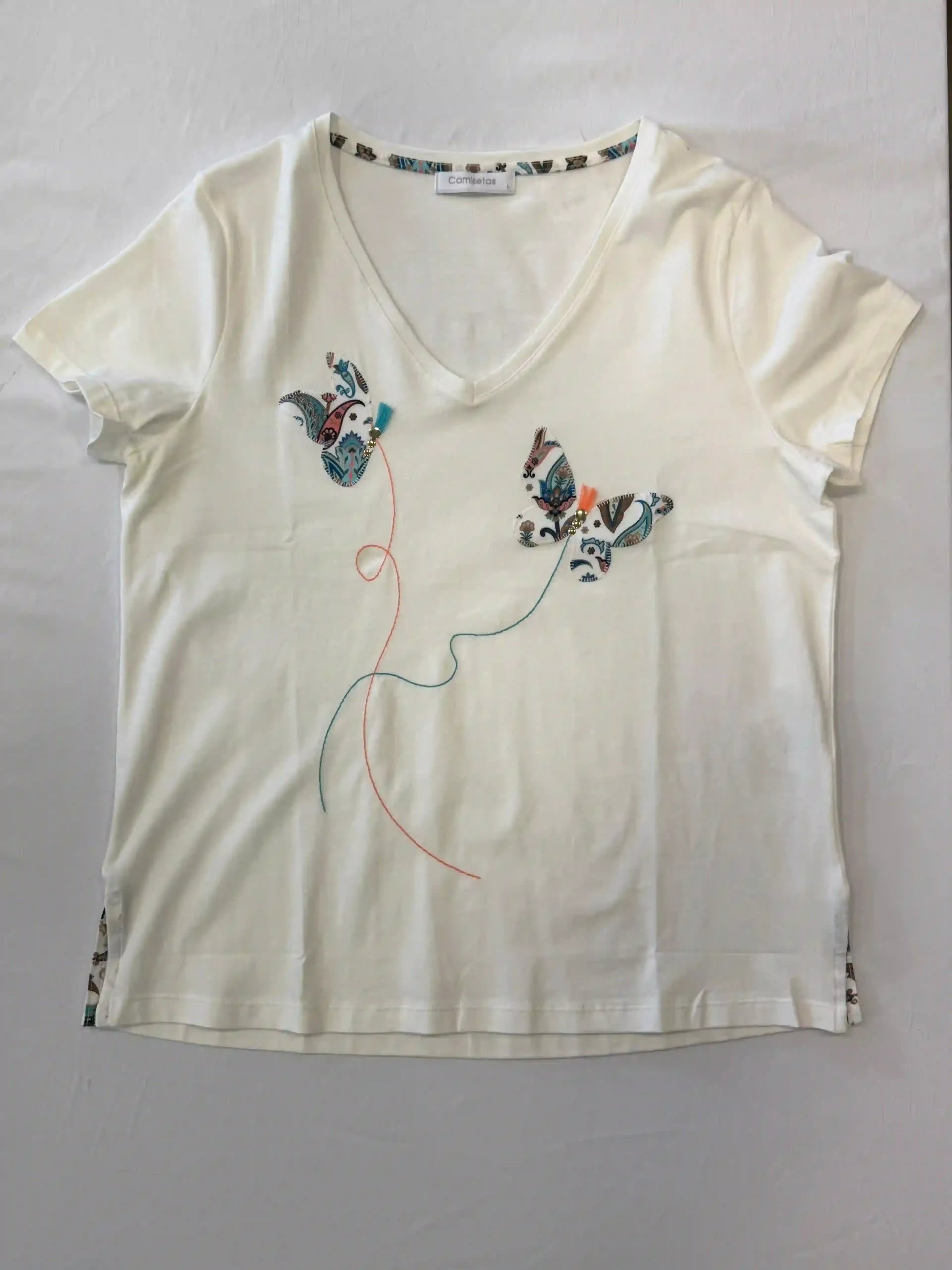 Camiseta color crema con cuello en V, mariposas florales bordadas en el pecho y un diseño colgante con cuentas.