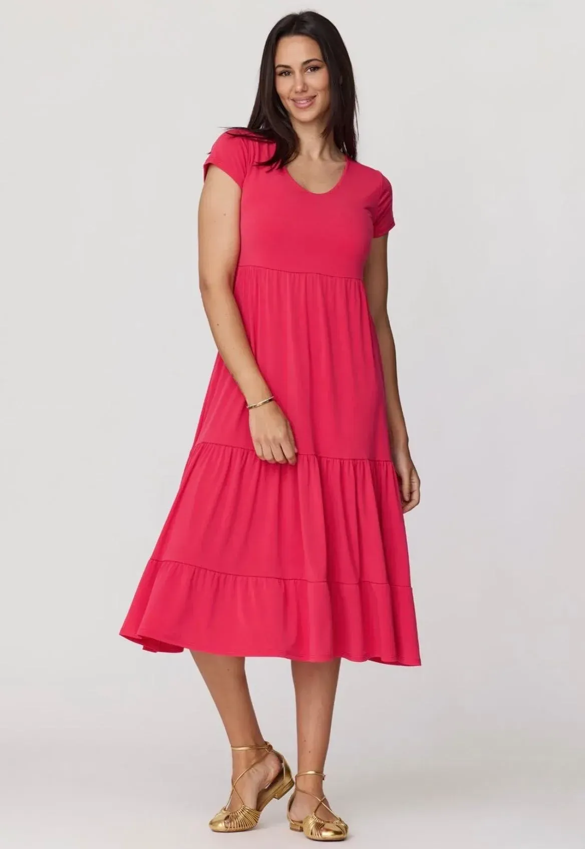 Mujer con un vestido rosa brillante y sandalias doradas, de pie frente a un fondo claro liso.