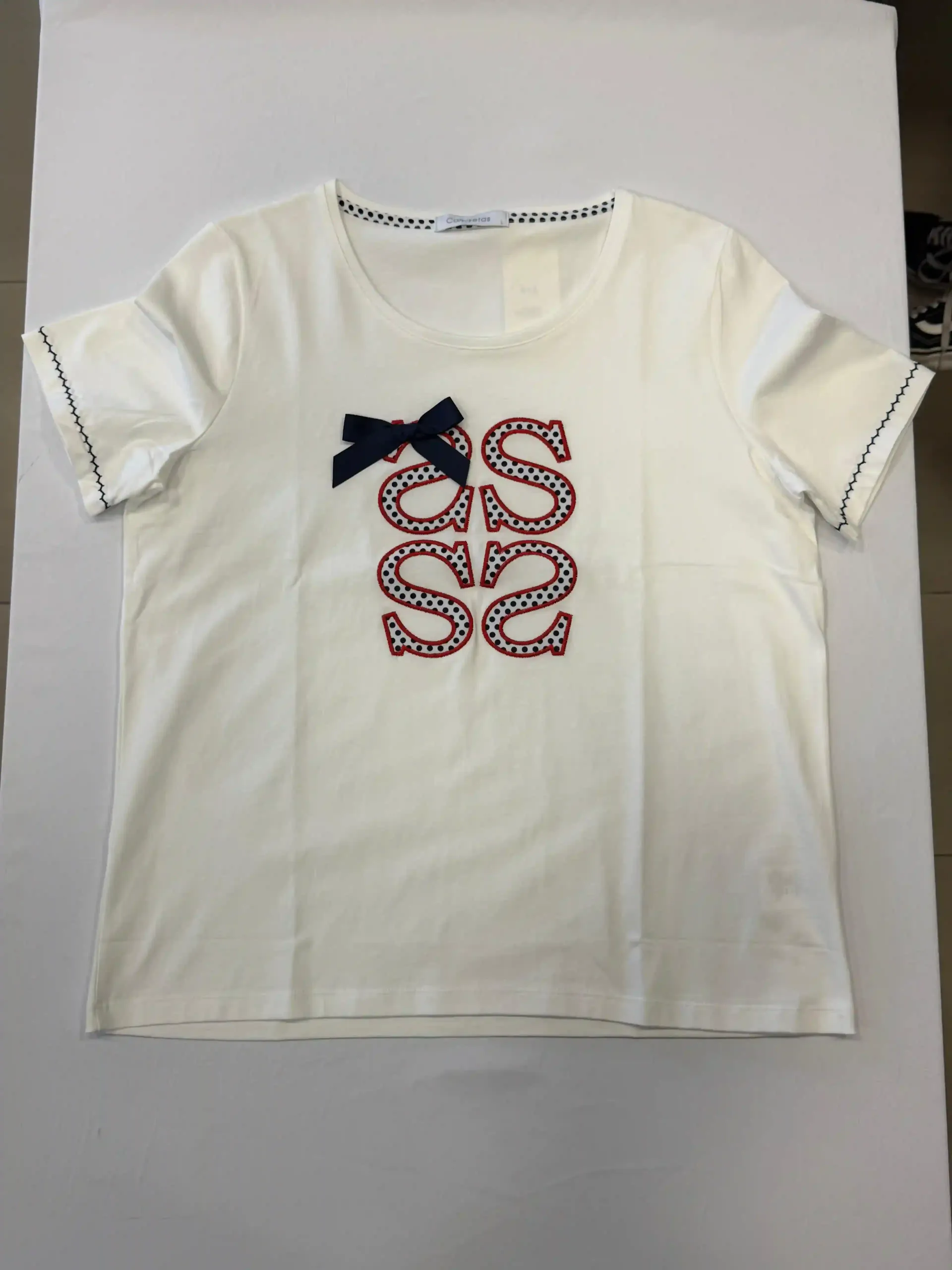 Camiseta blanca con un pequeño lazo azul marino y el logotipo “SS” estampado en rojo en el pecho.