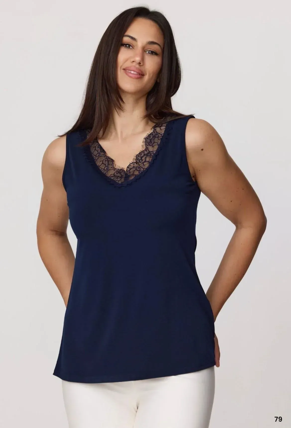 Mujer con una blusa azul marino sin mangas con escote de encaje y pantalones blancos, de pie sobre un fondo claro.