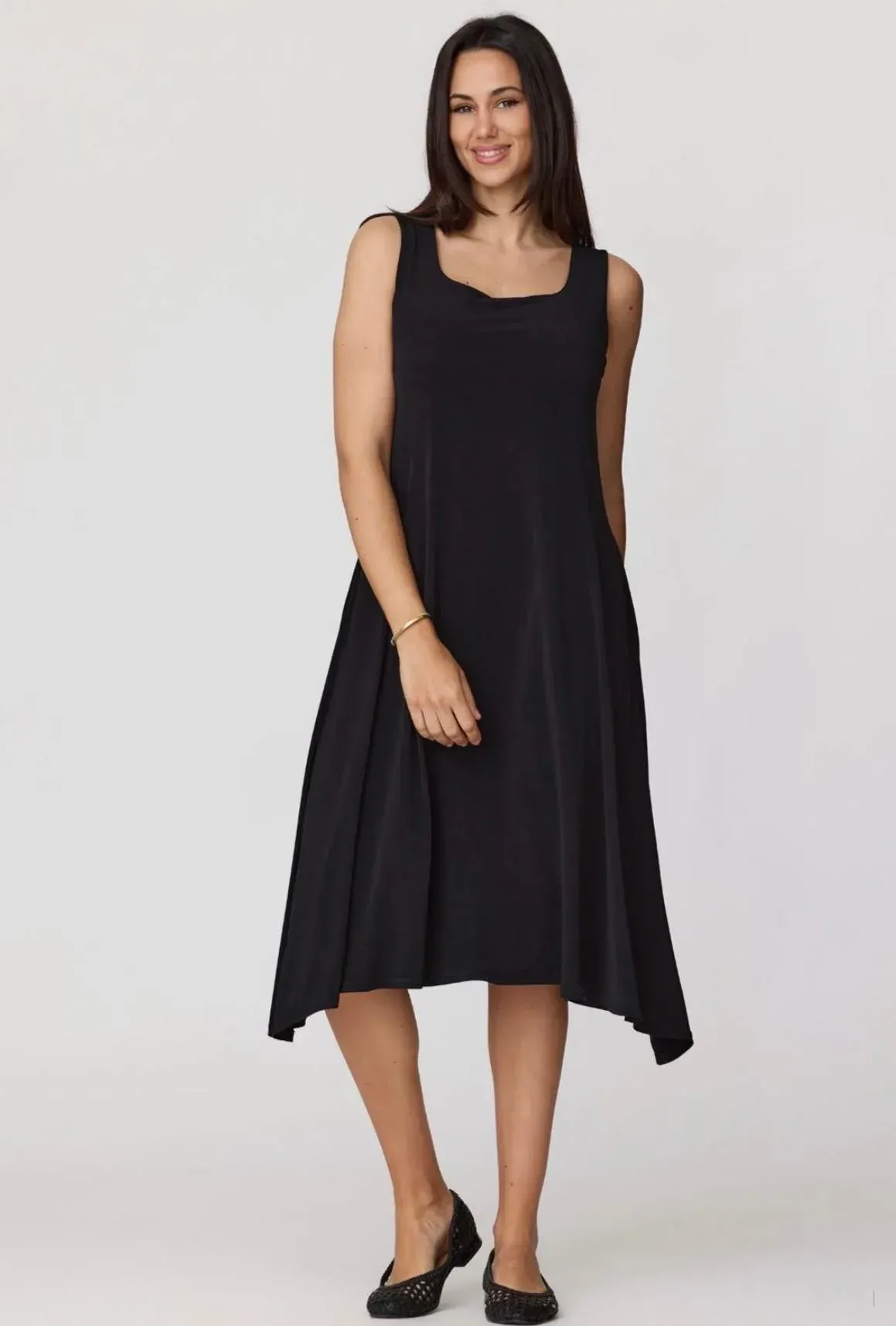 Mujer con vestido negro sin mangas y zapatos planos, de pie frente a un fondo claro liso.