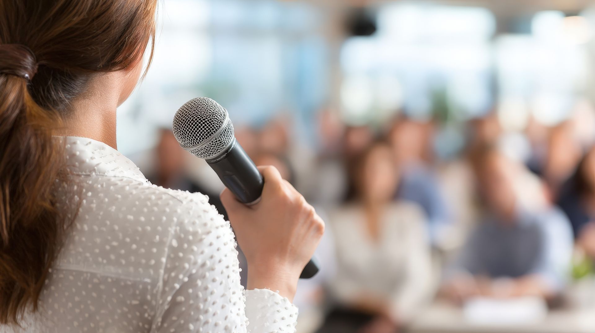 Une femme parle dans un microphone, s'adressant à un public flou dans un décor moderne.