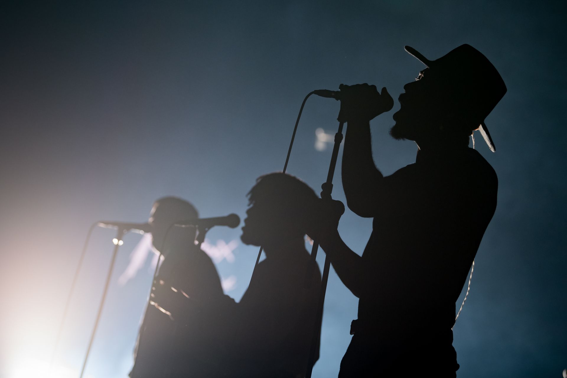 Des musiciens en silhouette chantent dans des microphones sur scène, rétroéclairés par les projecteurs.