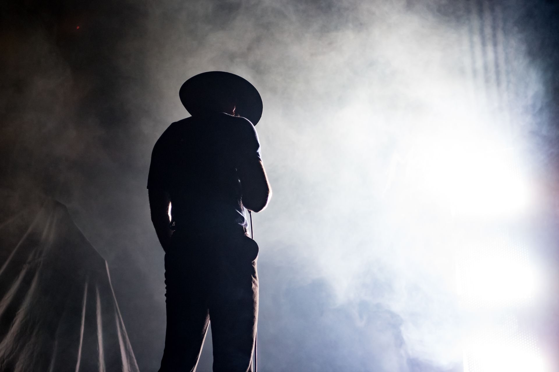 Silhouette d'une personne portant un chapeau, tenant un microphone, face à des lumières vives et à la fumée de scène.