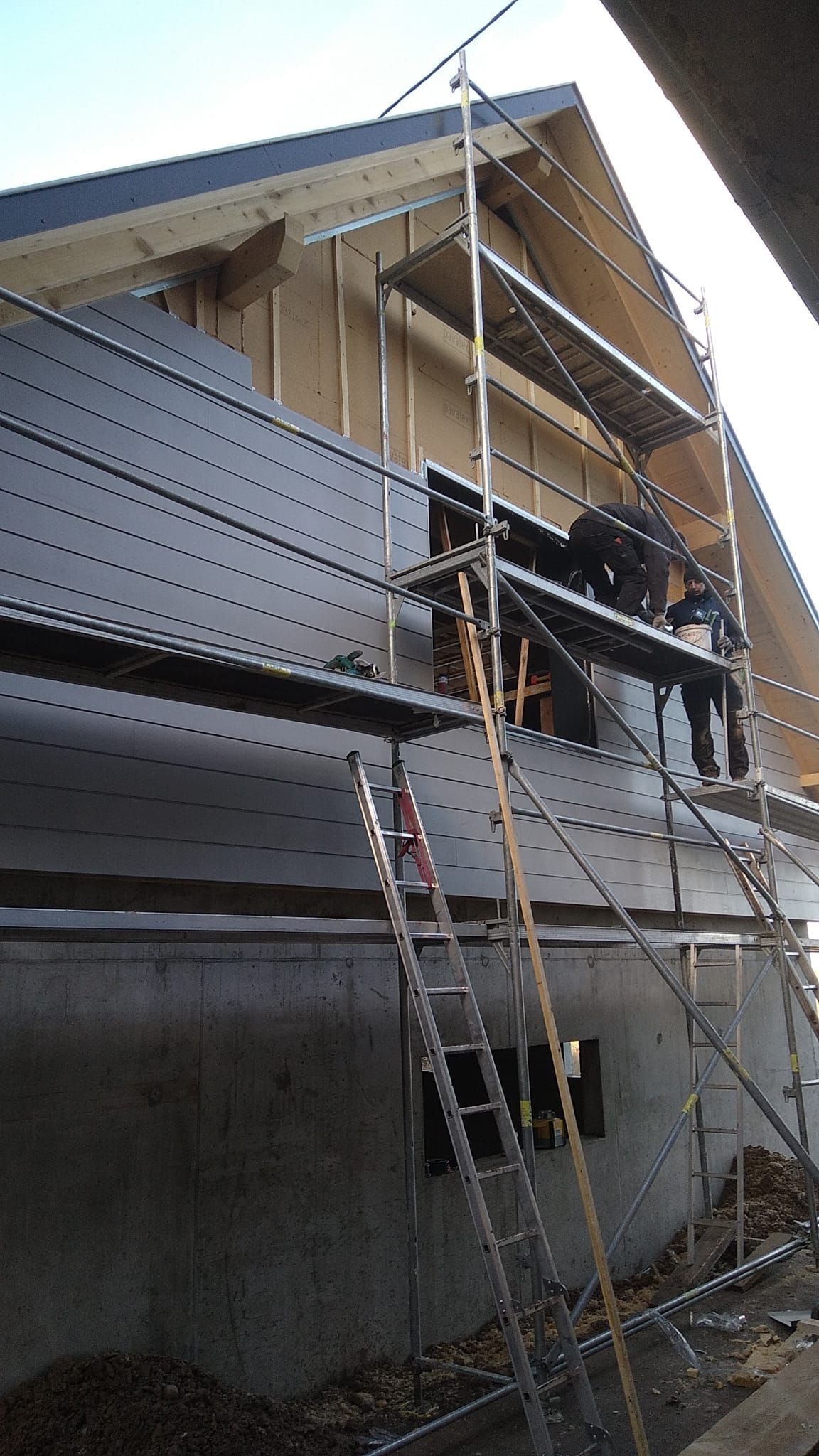 Des ouvriers du bâtiment posent un bardage noir sur un immeuble, à l'aide d'échafaudages et d'échelles.