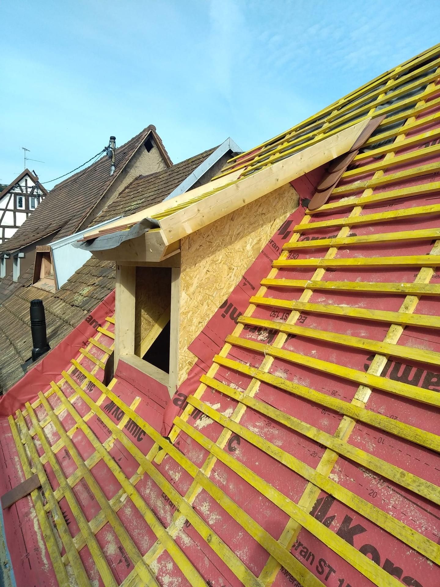 Construction de toiture : une lucarne encadrée de bois, entourée d'une sous-couche rouge et de chevrons jaunes.