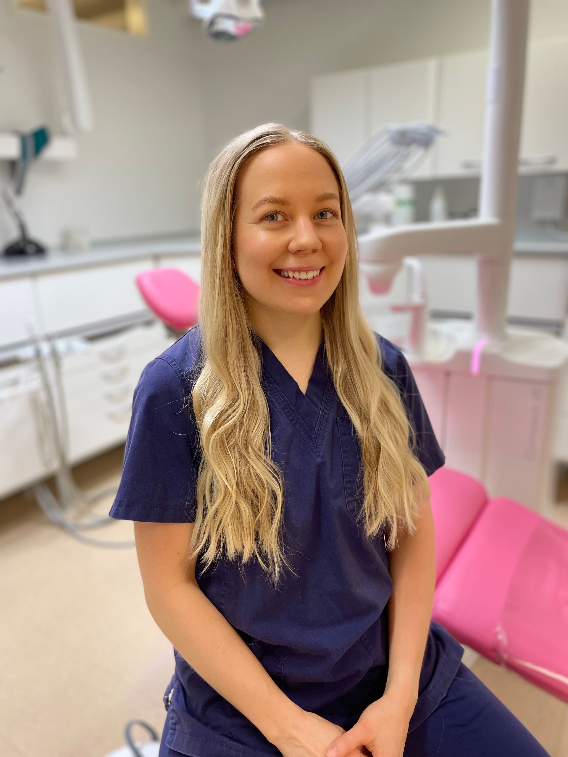 Hymyilevä hammaslääkäri tummansinisissä työvaatteissaan istuu kirkkaassa klinikassa, jossa on vaaleanpunaiset hoitotuolit
