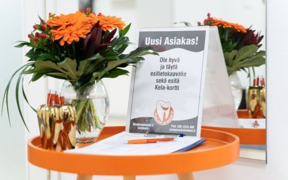 Oranssinvärisiä kukkia lasimaljakoissa ”Uusi Asiakas!” -kyltin vieressä oranssilla pöydällä.