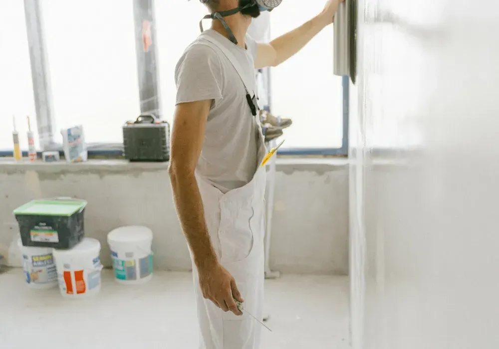Una persona con un mono blanco y un respirador pinta una pared en una habitación luminosa, con cubos de pintura cerca