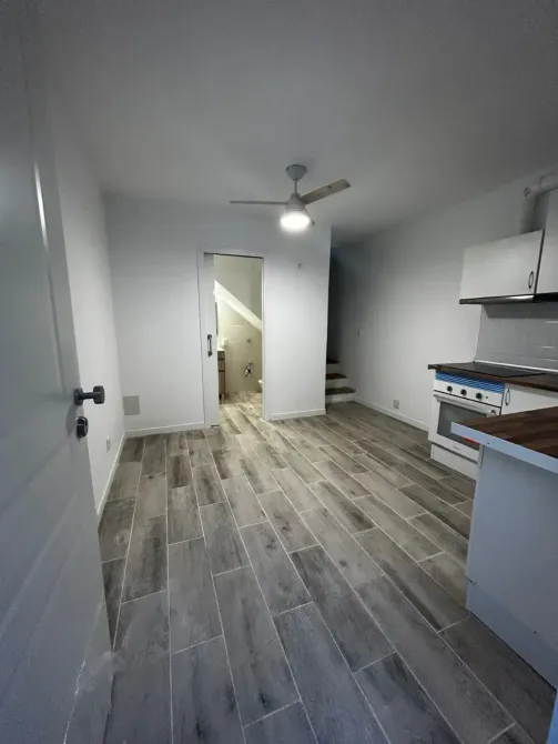 Cocina vacía con suelo de baldosas grises, paredes blancas, ventilador de techo y una puerta al fondo.