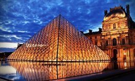 Una gran pirámide de cristal se ilumina por la noche frente a un edificio.