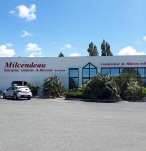 Bâtiment commercial avec une enseigne Milcendeau, une voiture garée et des arbustes sous un ciel bleu.