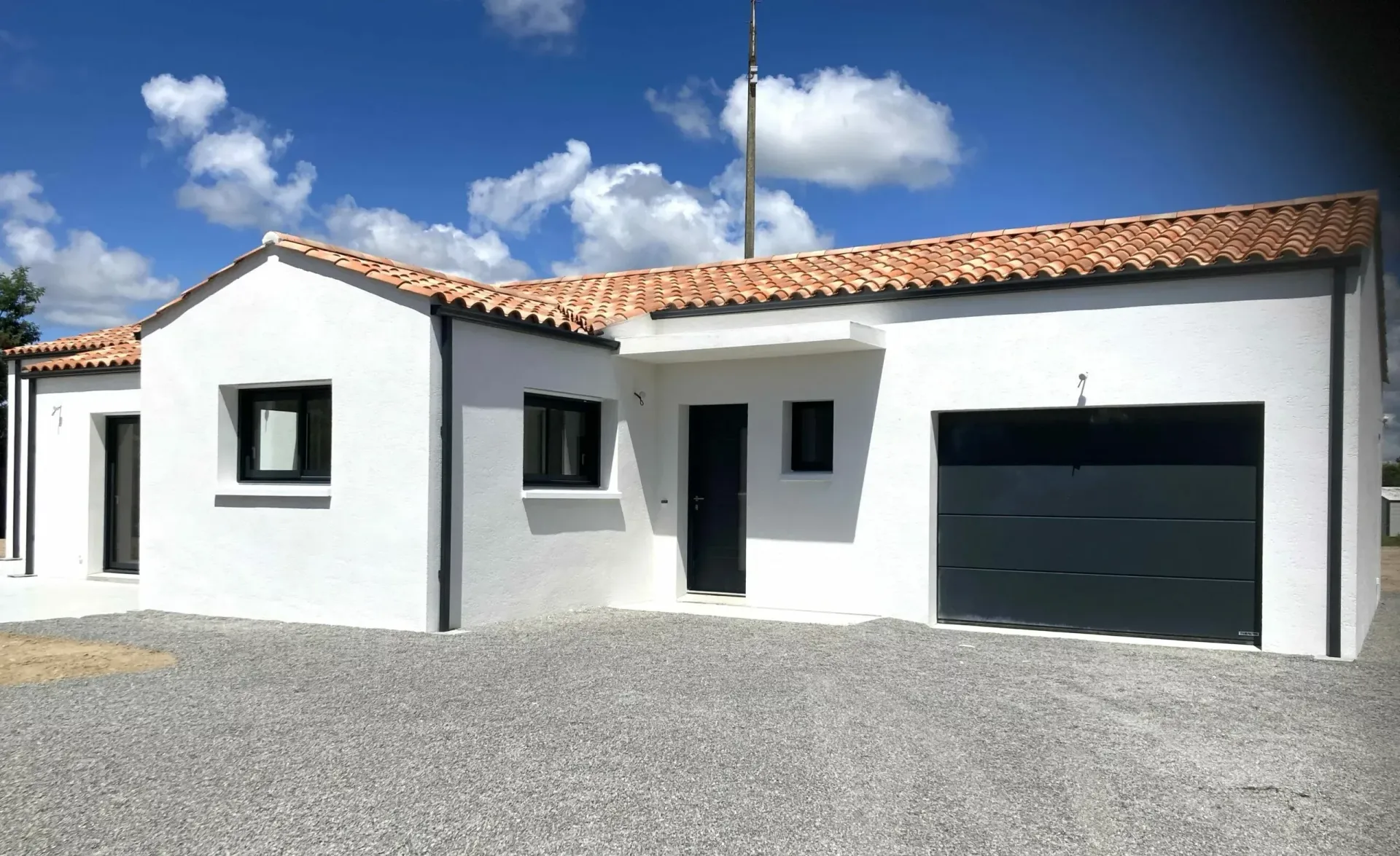 Construction Milcendeau : Maison blanche de plain-pied avec un toit en terre cuite, une porte de garage noire et une allée en gravier sous un ciel bleu.