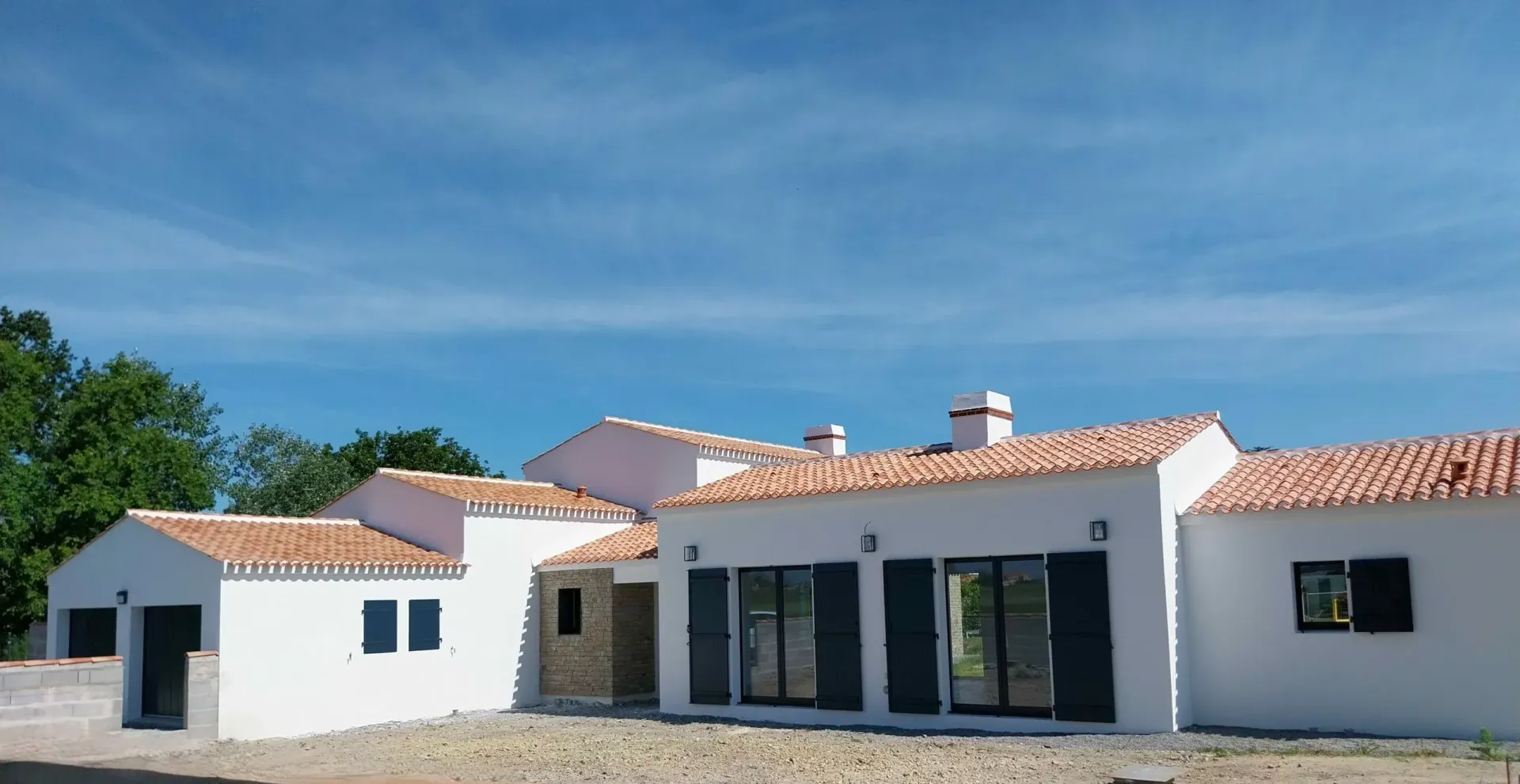 Construction Milcendeau : Maison moderne blanche avec des tuiles en terre cuite, des fenêtres à cadres noirs et une cour en gravier sous un ciel bleu.