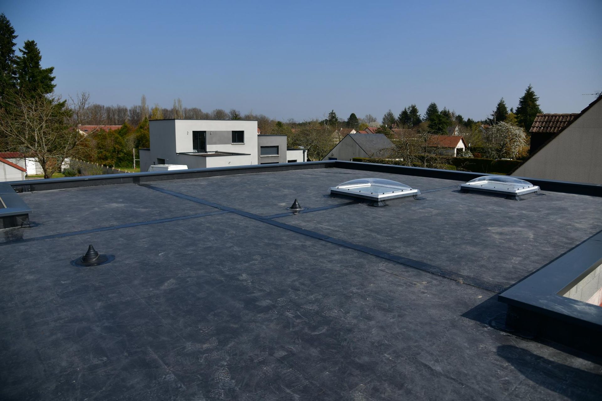 Toit plat avec des aérateurs et des puits de lumière, surplombant des maisons de banlieue sous un ciel bleu dégagé.