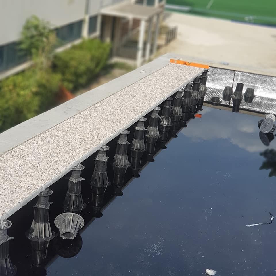Fontaine en carrelage noir avec plusieurs rangées de petits jets d’eau, à côté d’un muret en pierre, en extérieur