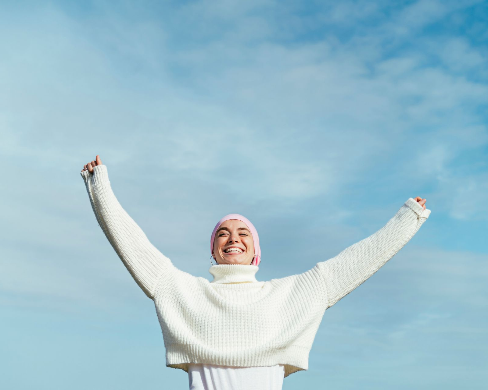 Femme en pull blanc et chapeau rose avec les bras levés, souriant et un ciel bleu.