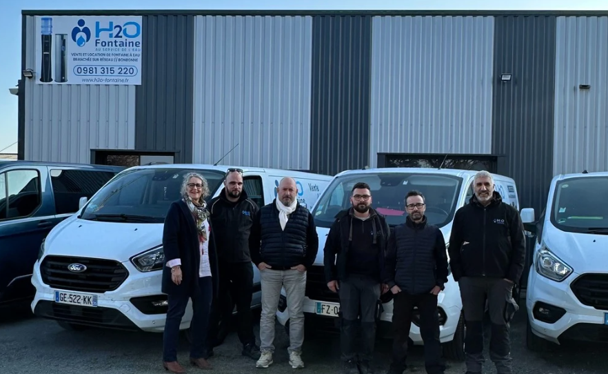Six personnes se tiennent devant des camionnettes blanches à l'extérieur d'un bâtiment industriel