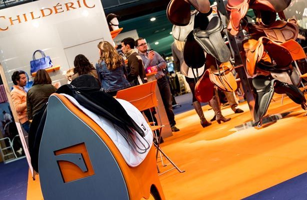 Un stand Childéric lors d'un salon professionnel, avec des selles en cuir exposées sur des présentoirs, un sol orange vif et des visiteurs qui flânent.