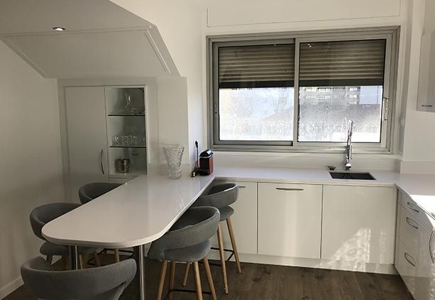 Une cuisine blanche moderne comprenant une table à manger en péninsule avec quatre tabourets gris, une fenêtre au-dessus de l'évier et un vaisselier.