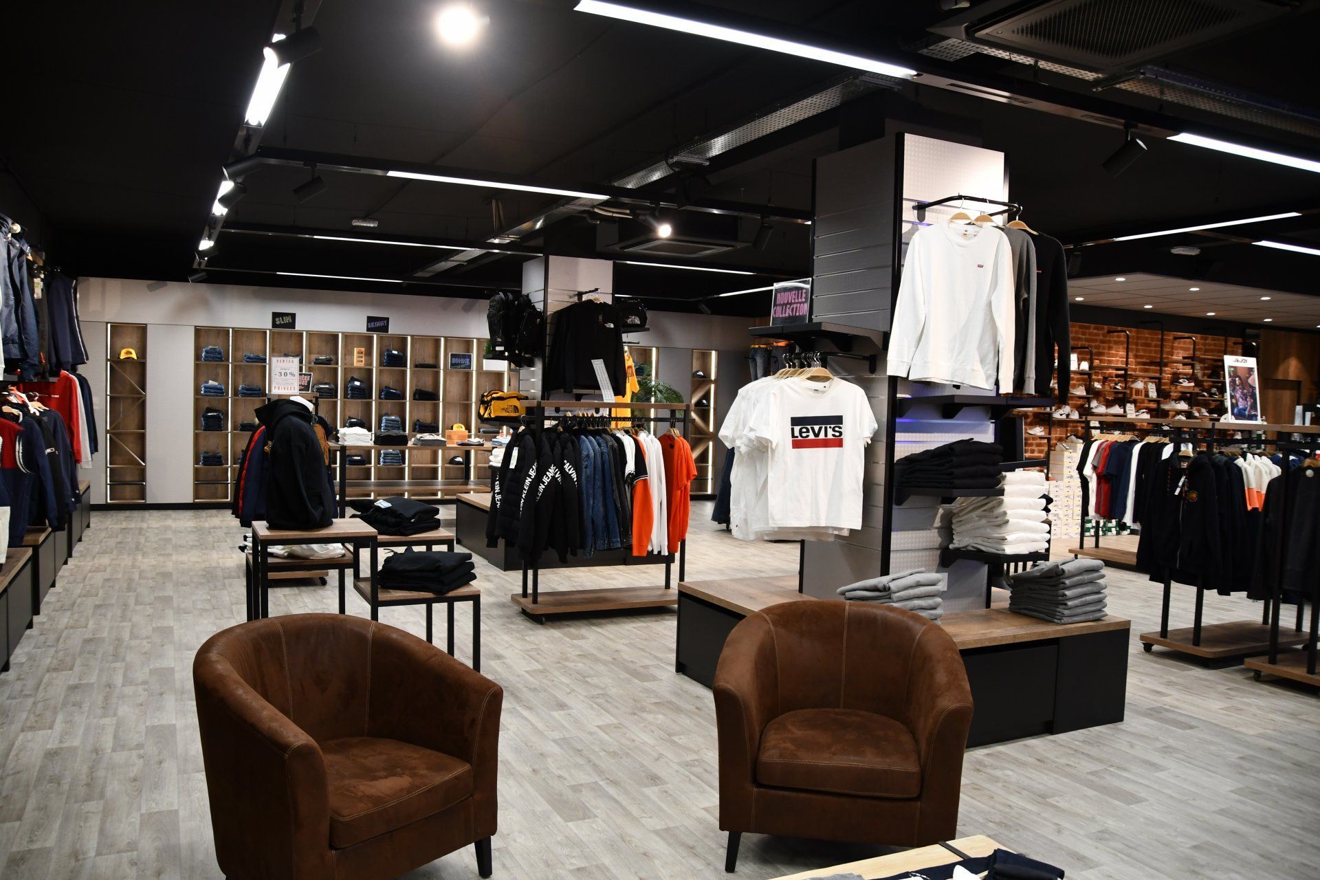 Intérieur d'un magasin de vêtements présentant des rangées de vêtements sur des portants et des étagères, avec deux fauteuils marron au premier plan.